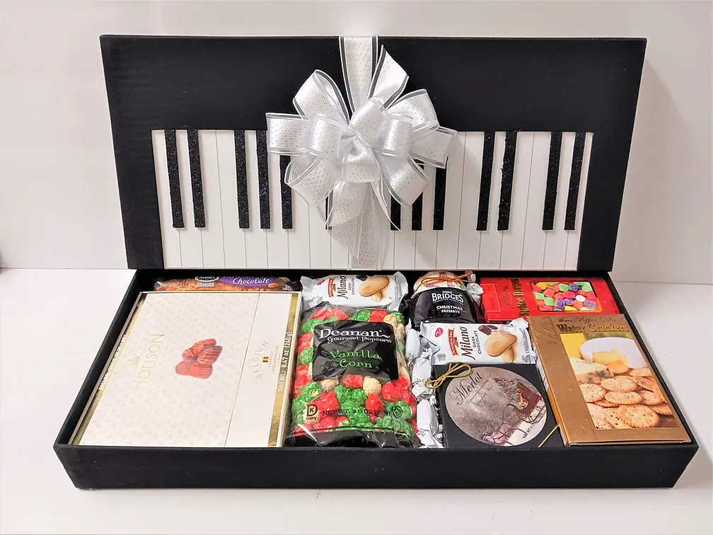 A gift box shaped like a piano, adorned with a white bow, filled with an assortment of snacks.